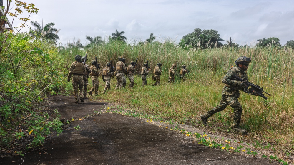 A group of armed military or special operations personnel are moving in formation along a paved path through a tropical or subtropical environment. The soldiers are equipped with tactical gear, body armor, helmets, and rifles. One soldier in the foreground is running while the rest advance in a column toward dense vegetation.