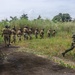 A group of armed military or special operations personnel are moving in formation along a paved path through a tropical or subtropical environment. The soldiers are equipped with tactical gear, body armor, helmets, and rifles. One soldier in the foreground is running while the rest advance in a column toward dense vegetation.