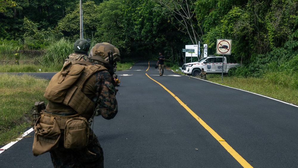 The image shows what appears to be a military training exercise on a paved road surrounded by dense tropical vegetation. Two armed soldiers in camouflage uniforms and tactical gear are in a forward-crouching position, with a civilian-clothed individual visible further down the road near a white vehicle. Signage in Spanish and a vehicle marked 'Zona 3 Aeronaut' with number '1017' suggest a Latin American location.