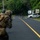 PANAMAX-26: V1/6, Partner forces conduct simulated air raid The image shows what appears to be a military training exercise on a paved road surrounded by dense tropical vegetation. Two armed soldiers in camouflage uniforms and tactical gear are in a forward-crouching position, with a civilian-clothed individual visible further down the road near a white vehicle. Signage in Spanish and a vehicle marked 'Zona 3 Aeronaut' with number '1017' suggest a Latin American location.