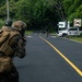 The image shows what appears to be a military training exercise on a paved road surrounded by dense tropical vegetation. Two armed soldiers in camouflage uniforms and tactical gear are in a forward-crouching position, with a civilian-clothed individual visible further down the road near a white vehicle. Signage in Spanish and a vehicle marked 'Zona 3 Aeronaut' with number '1017' suggest a Latin American location.