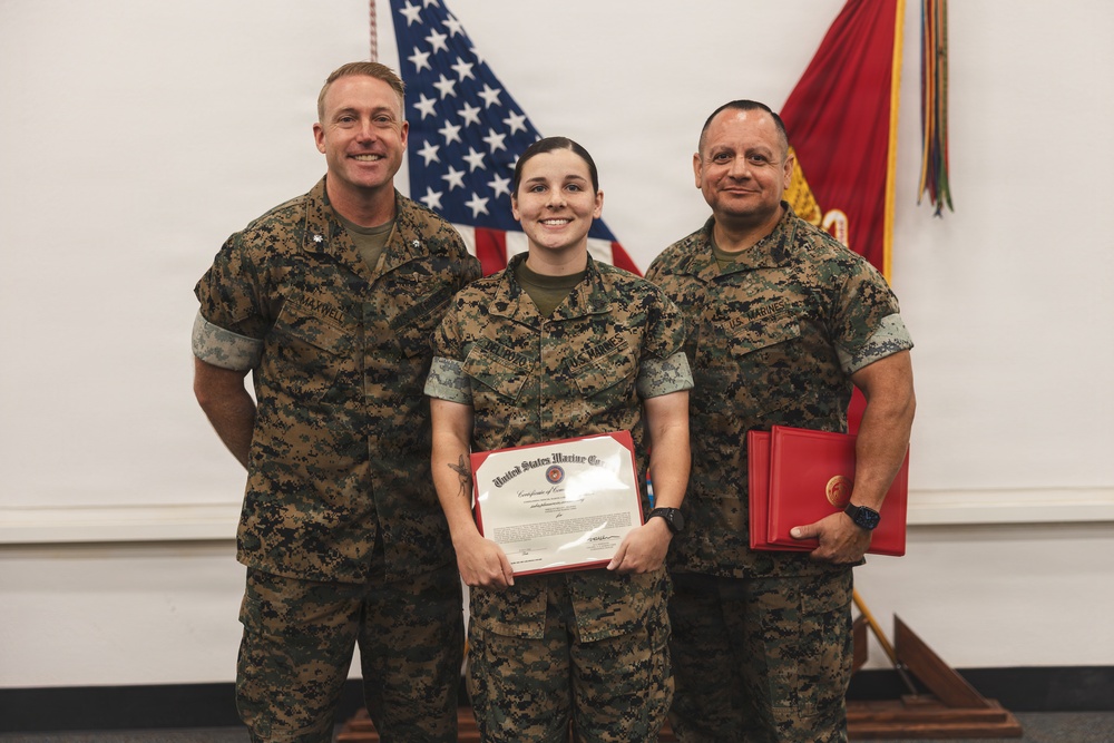 Three United States Marines in digital camouflage (MARPAT) uniforms pose for a formal photo during what appears to be an awards or recognition ceremony. The central figure, a female Marine, is holding a United States Marine Corps Certificate of Commendation, while the Marine on the right holds red presentation folders. An American flag and a Marine Corps flag are visible in the background.