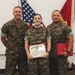 Three United States Marines in digital camouflage (MARPAT) uniforms pose for a formal photo during what appears to be an awards or recognition ceremony. The central figure, a female Marine, is holding a United States Marine Corps Certificate of Commendation, while the Marine on the right holds red presentation folders. An American flag and a Marine Corps flag are visible in the background.