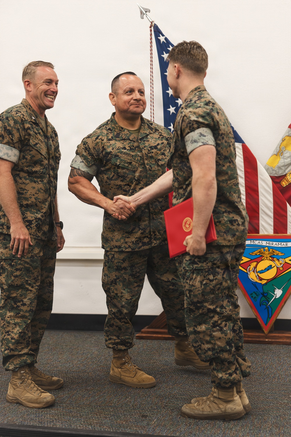 Three U.S. Marines in MARPAT woodland camouflage uniforms are participating in a formal military ceremony at MCAS Miramar. The central figure, whose name tape reads 'GARCIA,' is shaking hands with a younger Marine who is holding a red award folder bearing the Marine Corps emblem. A third Marine, whose name tape appears to read 'MAYFIELD,' stands to the left smiling broadly.