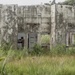 The image shows an abandoned, deteriorating concrete structure in a tropical environment overgrown with tall grass and vegetation. A single armed and helmeted individual in military-style camouflage gear is visible in the doorway of the structure, appearing to conduct a tactical entry or clearing operation. The building shows significant weathering, moss growth, and structural decay consistent with long-term abandonment.
