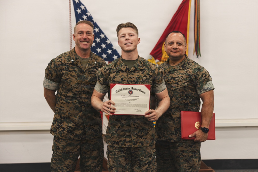 Three United States Marine Corps personnel pose together during a formal recognition ceremony. The central figure holds a 'Certificate of Commendation' issued by the United States Marine Corps, while the individual on the right holds a red presentation folder. All three are wearing MARPAT (Marine Pattern) camouflage uniforms.