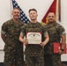 Three United States Marine Corps personnel pose together during a formal recognition ceremony. The central figure holds a 'Certificate of Commendation' issued by the United States Marine Corps, while the individual on the right holds a red presentation folder. All three are wearing MARPAT (Marine Pattern) camouflage uniforms.