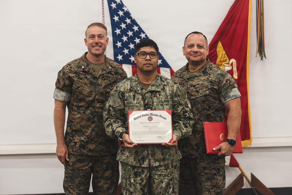 Three military personnel pose for a formal photo during an award ceremony. The center individual, wearing U.S. Navy NWU (Navy Working Uniform), holds a United States Marine Corps Certificate of Commendation. Two Marines in MARPAT camouflage flank the recipient, with one holding a red presentation folder.