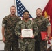 Three military personnel pose for a formal photo during an award ceremony. The center individual, wearing U.S. Navy NWU (Navy Working Uniform), holds a United States Marine Corps Certificate of Commendation. Two Marines in MARPAT camouflage flank the recipient, with one holding a red presentation folder.