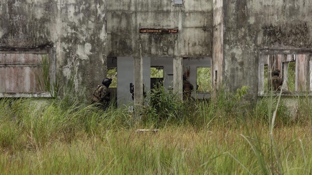 The image shows multiple armed personnel in camouflage uniforms conducting what appears to be a tactical training exercise or clearing operation at an abandoned, deteriorating concrete structure. The building shows significant weathering, vegetation overgrowth, and structural decay. Personnel are positioned at entry points and along the exterior wall of the structure.