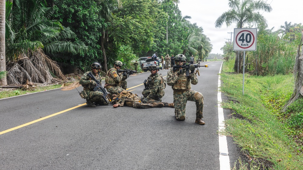 A group of armed military or special operations personnel are positioned on a paved road in a tropical environment, appearing to conduct a tactical exercise or operation. Several soldiers are in kneeling and prone positions with weapons raised, while one individual appears to be lying on the ground. A speed limit sign reading 40 Km/h is visible on the right side of the road.