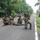 PANAMAX-26: V1/6, Partner forces conduct simulated air raid A group of armed military or special operations personnel are positioned on a paved road in a tropical environment, appearing to conduct a tactical exercise or operation. Several soldiers are in kneeling and prone positions with weapons raised, while one individual appears to be lying on the ground. A speed limit sign reading 40 Km/h is visible on the right side of the road.