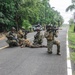 A group of armed military or special operations personnel are positioned on a paved road in a tropical environment, appearing to conduct a tactical exercise or operation. Several soldiers are in kneeling and prone positions with weapons raised, while one individual appears to be lying on the ground. A speed limit sign reading 40 Km/h is visible on the right side of the road.