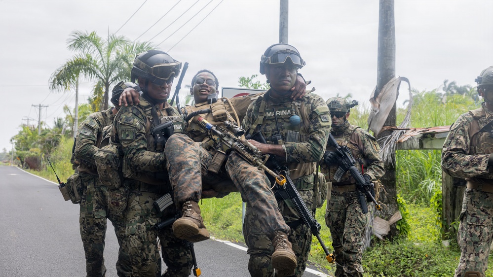 Military personnel in camouflage uniforms are conducting what appears to be a casualty evacuation drill or exercise on a road in a tropical environment. Two soldiers bearing 'MARINA' insignia are carrying a third soldier who is smiling, suggesting a training scenario rather than a real emergency. Additional armed personnel are visible in the background.