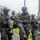 PANAMAX-26: V1/6, Partner forces conduct simulated air raid Military personnel in camouflage uniforms are conducting what appears to be a casualty evacuation drill or exercise on a road in a tropical environment. Two soldiers bearing 'MARINA' insignia are carrying a third soldier who is smiling, suggesting a training scenario rather than a real emergency. Additional armed personnel are visible in the background.