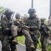 Military personnel in camouflage uniforms are conducting what appears to be a casualty evacuation drill or exercise on a road in a tropical environment. Two soldiers bearing 'MARINA' insignia are carrying a third soldier who is smiling, suggesting a training scenario rather than a real emergency. Additional armed personnel are visible in the background.