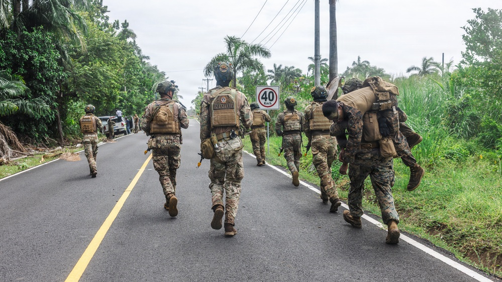 A group of military personnel in camouflage uniforms and tactical gear are moving along a paved road flanked by tropical vegetation. One soldier on the right is carrying another person on their back, suggesting a casualty evacuation drill or training exercise. A speed limit sign reading 40 km/h is visible along the roadside.