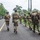 PANAMAX-26: V1/6, Partner forces conduct simulated air raid A group of military personnel in camouflage uniforms and tactical gear are moving along a paved road flanked by tropical vegetation. One soldier on the right is carrying another person on their back, suggesting a casualty evacuation drill or training exercise. A speed limit sign reading 40 km/h is visible along the roadside.