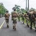 A group of military personnel in camouflage uniforms and tactical gear are moving along a paved road flanked by tropical vegetation. One soldier on the right is carrying another person on their back, suggesting a casualty evacuation drill or training exercise. A speed limit sign reading 40 km/h is visible along the roadside.