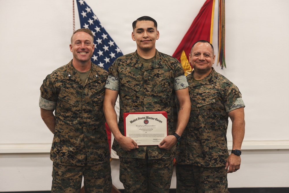 Three United States Marines in digital camouflage (MARPAT) uniforms are posed together for a formal photo. The central figure is holding a United States Marine Corps Certificate of Commendation with a red border. The setting appears to be an indoor military ceremony room with an American flag and a Marine Corps flag visible in the background.