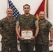 Three United States Marines in digital camouflage (MARPAT) uniforms are posed together for a formal photo. The central figure is holding a United States Marine Corps Certificate of Commendation with a red border. The setting appears to be an indoor military ceremony room with an American flag and a Marine Corps flag visible in the background.