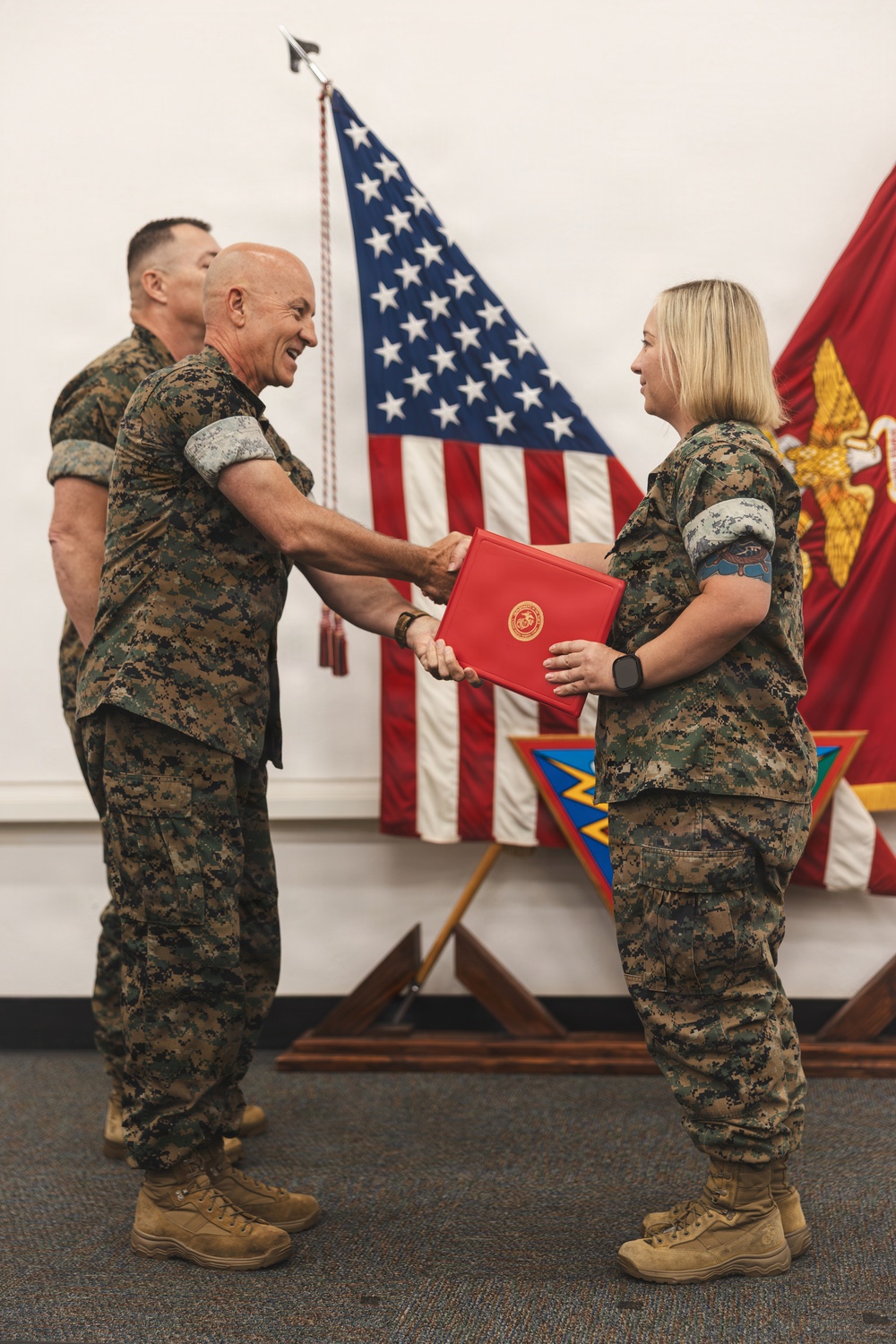 A military ceremony is taking place in which a senior Marine officer is presenting a red official folder to a female Marine while shaking her hand. Both individuals are wearing USMC MARPAT woodland camouflage uniforms. The setting includes an American flag and a Marine Corps flag displayed in the background.