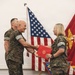 A military ceremony is taking place in which a senior Marine officer is presenting a red official folder to a female Marine while shaking her hand. Both individuals are wearing USMC MARPAT woodland camouflage uniforms. The setting includes an American flag and a Marine Corps flag displayed in the background.