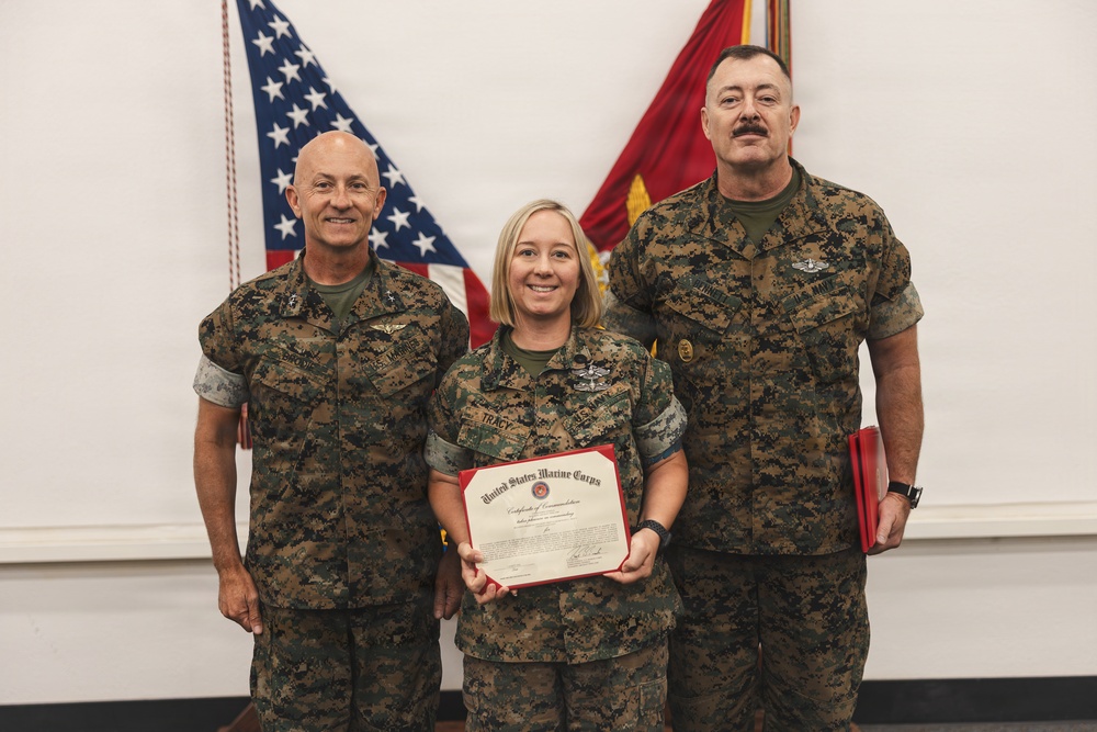 Three United States Marine Corps personnel are posed together during a formal recognition ceremony. The central figure, a female Marine with the name tape 'TRACY' visible, is holding a United States Marine Corps Certificate of Commendation. Two male Marines flank her, with an American flag and Marine Corps colors visible in the background.