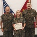 Three United States Marine Corps personnel are posed together during a formal recognition ceremony. The central figure, a female Marine with the name tape 'TRACY' visible, is holding a United States Marine Corps Certificate of Commendation. Two male Marines flank her, with an American flag and Marine Corps colors visible in the background.