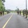 PANAMAX-26: V1/6, Partner forces conduct simulated air raid A group of armed military or law enforcement personnel in full tactical gear are conducting a patrol or movement along a paved two-lane road in a tropical environment. The personnel are spread across both sides of the road, moving away from the camera in a column formation. The setting appears to be a rural or semi-rural area with palm trees, lush vegetation, and buildings visible in the distance.
