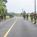 A group of armed military or law enforcement personnel in full tactical gear are conducting a patrol or movement along a paved two-lane road in a tropical environment. The personnel are spread across both sides of the road, moving away from the camera in a column formation. The setting appears to be a rural or semi-rural area with palm trees, lush vegetation, and buildings visible in the distance.