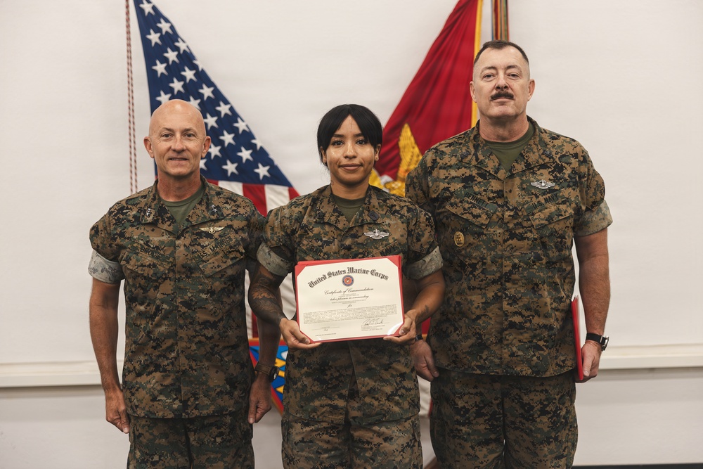 Three United States Marine Corps personnel are posed for a formal recognition photograph. The central figure, a female Marine, is holding a United States Marine Corps Certificate of Commendation. Two senior Marines flank her on either side against a backdrop of American and USMC flags.