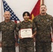 Three United States Marine Corps personnel are posed for a formal recognition photograph. The central figure, a female Marine, is holding a United States Marine Corps Certificate of Commendation. Two senior Marines flank her on either side against a backdrop of American and USMC flags.