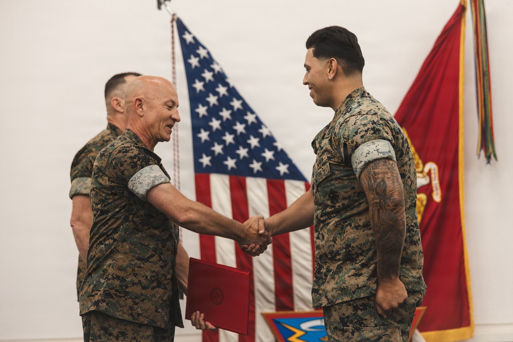 Two U.S. Marines in MARPAT digital camouflage uniforms are shaking hands during what appears to be a formal military ceremony. The senior Marine on the left is holding a red presentation folder, suggesting an award or commendation is being presented. A third Marine is visible in the background, and both the U.S. flag and a Marine Corps unit colors flag are displayed behind them.