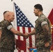 Two U.S. Marines in MARPAT digital camouflage uniforms are shaking hands during what appears to be a formal military ceremony. The senior Marine on the left is holding a red presentation folder, suggesting an award or commendation is being presented. A third Marine is visible in the background, and both the U.S. flag and a Marine Corps unit colors flag are displayed behind them.
