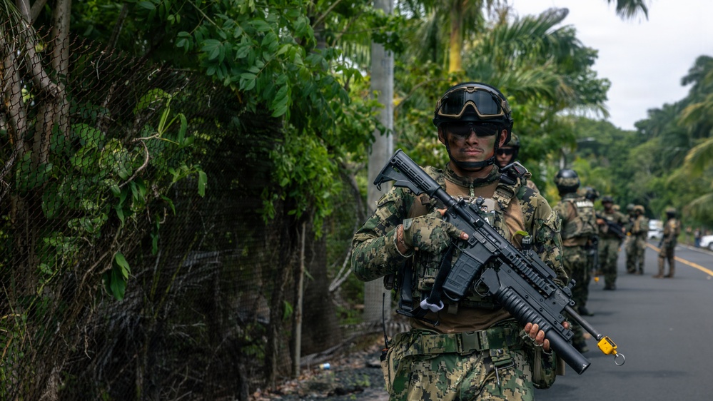 A column of armed military or special operations personnel is patrolling along a paved road bordered by dense tropical vegetation and a chain-link fence. The lead soldier in the foreground is in full tactical gear, carrying an assault rifle with an underbarrel attachment. The tropical environment and vegetation suggest a Central or South American or Southeast Asian setting.