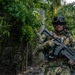 A column of armed military or special operations personnel is patrolling along a paved road bordered by dense tropical vegetation and a chain-link fence. The lead soldier in the foreground is in full tactical gear, carrying an assault rifle with an underbarrel attachment. The tropical environment and vegetation suggest a Central or South American or Southeast Asian setting.