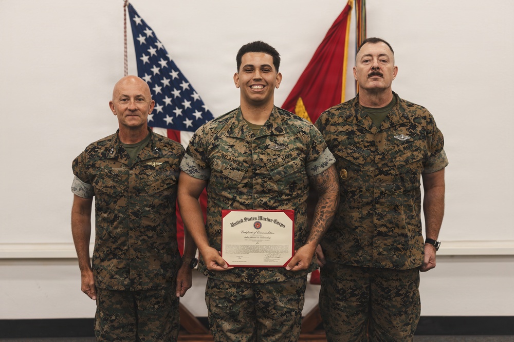 Three military personnel in MARPAT camouflage uniforms pose for a formal photograph. The central figure holds a United States Marine Corps Certificate of Commendation in a red presentation folder. Two flags — the American flag and a red Marine Corps flag — are visible in the background.