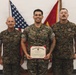Three military personnel in MARPAT camouflage uniforms pose for a formal photograph. The central figure holds a United States Marine Corps Certificate of Commendation in a red presentation folder. Two flags — the American flag and a red Marine Corps flag — are visible in the background.
