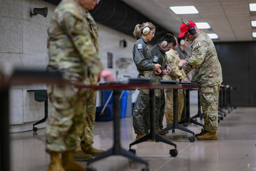 Military personnel are gathered at an indoor shooting range, standing at individual shooting benches. Multiple individuals wearing hearing protection are handling firearms or equipment at the tables. The group includes personnel in both OCP (Operational Camouflage Pattern) uniforms and flight suits.
