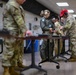 Military personnel are gathered at an indoor shooting range, standing at individual shooting benches. Multiple individuals wearing hearing protection are handling firearms or equipment at the tables. The group includes personnel in both OCP (Operational Camouflage Pattern) uniforms and flight suits.