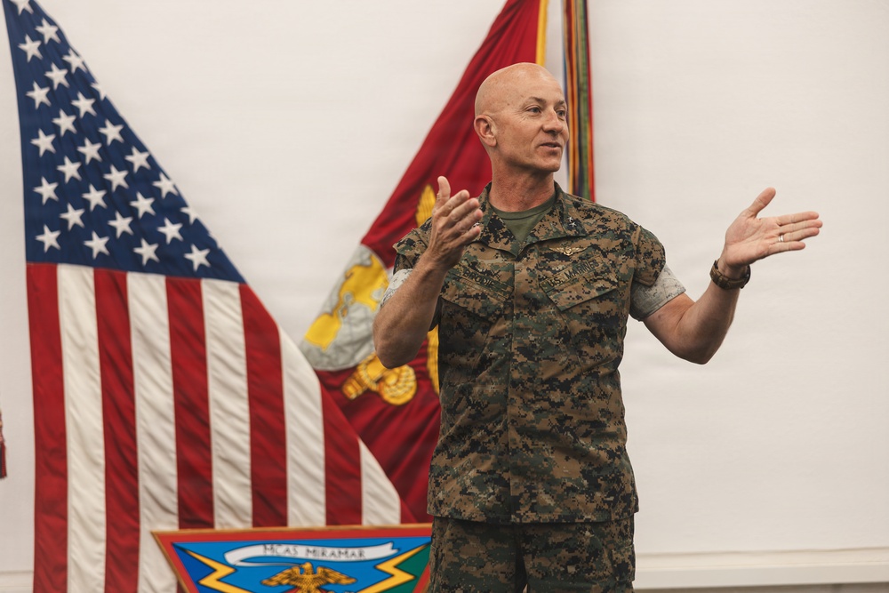 A U.S. Marine Corps officer in MARPAT camouflage uniform is speaking or presenting at an official military ceremony or briefing. He is gesturing with both hands in an expressive manner, standing in front of multiple flags including the U.S. national flag and a U.S. Marine Corps flag. A unit shield bearing the text 'MCAS Miramar' is visible in the lower foreground.