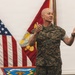 A U.S. Marine Corps officer in MARPAT camouflage uniform is speaking or presenting at an official military ceremony or briefing. He is gesturing with both hands in an expressive manner, standing in front of multiple flags including the U.S. national flag and a U.S. Marine Corps flag. A unit shield bearing the text 'MCAS Miramar' is visible in the lower foreground.