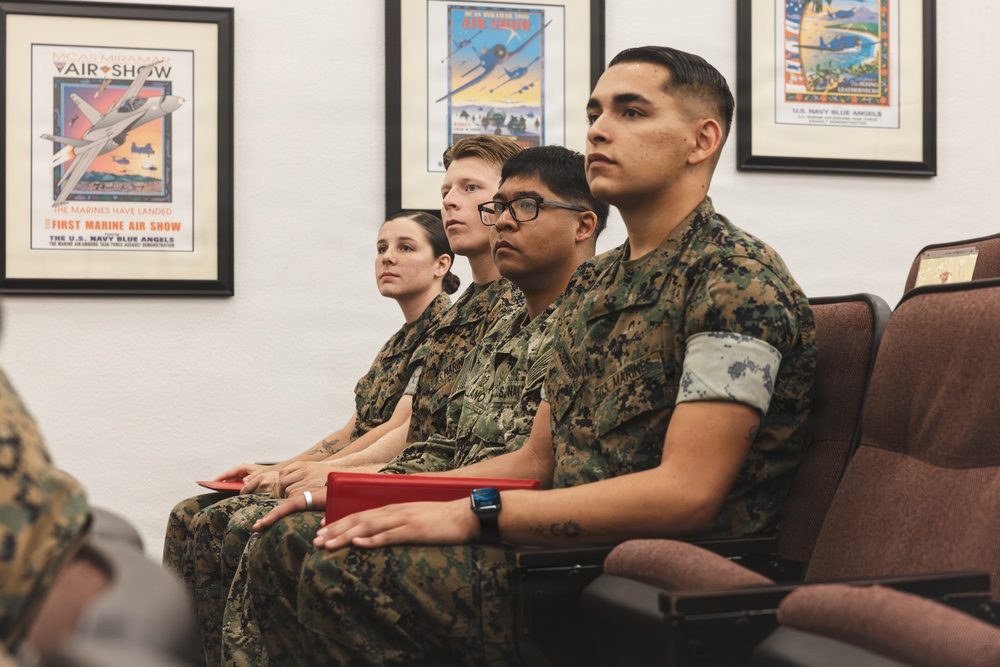 Four U.S. Marines in woodland MARPAT camouflage uniforms are seated in chairs, appearing to attend a briefing or presentation. They are in an indoor room decorated with framed military aviation posters, including references to the U.S. Navy Blue Angels and Marine Air Shows. The Marines appear attentive and focused on something outside the frame.