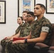 Four U.S. Marines in woodland MARPAT camouflage uniforms are seated in chairs, appearing to attend a briefing or presentation. They are in an indoor room decorated with framed military aviation posters, including references to the U.S. Navy Blue Angels and Marine Air Shows. The Marines appear attentive and focused on something outside the frame.