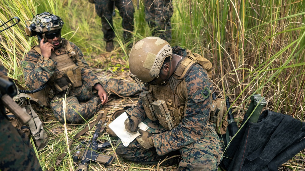 Two military personnel in camouflage uniforms are positioned in a grassy field environment, engaged in tactical communications and documentation activities. The foreground soldier is writing on a notepad while wearing full combat gear including a tan helmet, plate carrier, and magazine pouches. A second soldier in the background is using a radio handset, suggesting coordinated field operations or training exercise.