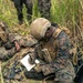 Two military personnel in camouflage uniforms are positioned in a grassy field environment, engaged in tactical communications and documentation activities. The foreground soldier is writing on a notepad while wearing full combat gear including a tan helmet, plate carrier, and magazine pouches. A second soldier in the background is using a radio handset, suggesting coordinated field operations or training exercise.