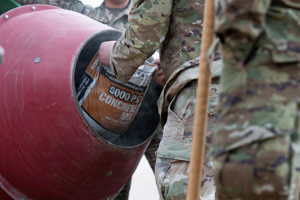 Military personnel in camouflage uniforms are pouring a bag of 5000 PSI concrete mix into a red cement mixer drum. The activity appears to be a construction or engineering task being performed by soldiers, likely during a training exercise or field operation. At least two individuals in OCP (Operational Camouflage Pattern) uniforms are visible.