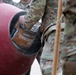 Military personnel in camouflage uniforms are pouring a bag of 5000 PSI concrete mix into a red cement mixer drum. The activity appears to be a construction or engineering task being performed by soldiers, likely during a training exercise or field operation. At least two individuals in OCP (Operational Camouflage Pattern) uniforms are visible.