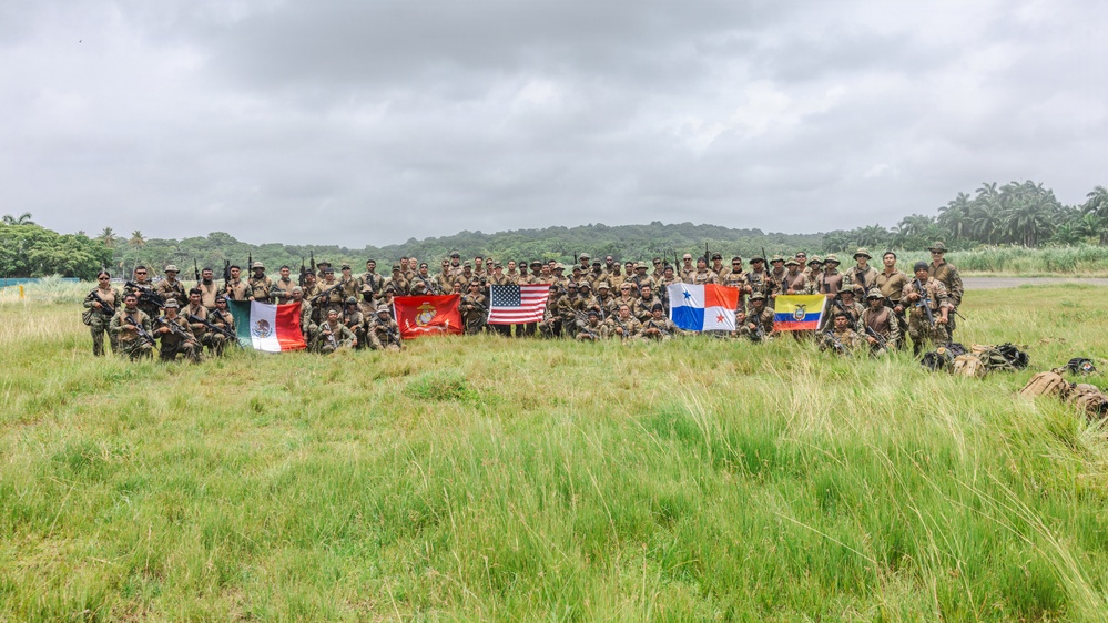 A large group of military personnel from multiple nations pose for a group photograph in an open grassy field with tropical vegetation in the background. Participants are holding several national and military flags, indicating a multinational military exercise or joint training event. The flags visible include those of Mexico, the United States, Panama, Ecuador, and a U.S. Marine Corps flag.